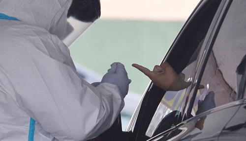 Photograph of a person wearing protective gear standing by a car with a rolled-down window. An individual within the car is holding their finger out of the window so that a pinprick of blood can be taken for an antibody test.