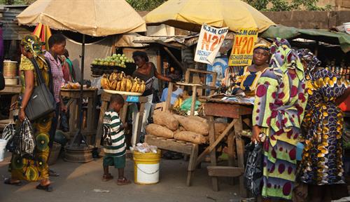 A scene from a street market in Lagos, Nigeria, the largest city in Africa. Several stalls selling produce are shown, as well as vendors and shoppers.