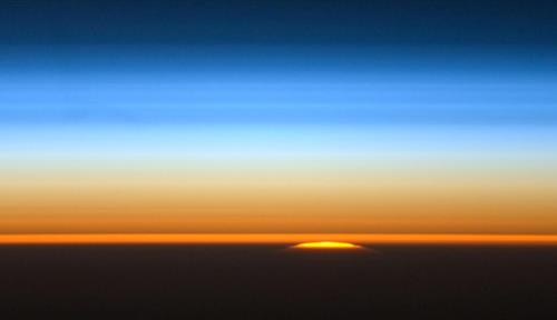 Photo taken by the crew of the International Space Station of the sun peeking over the edge of the planet showing a sky that darkens as you move away from Earth.