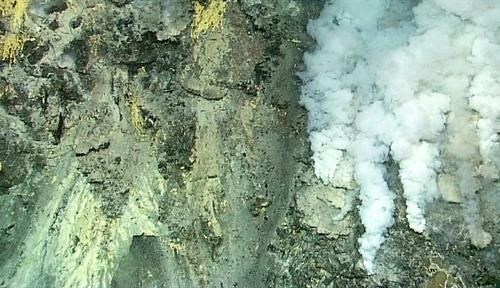 Photograph of a volcanic vent on the ocean floor. Colored minerals can be seen on the rock, as well as plumes of white smoke.