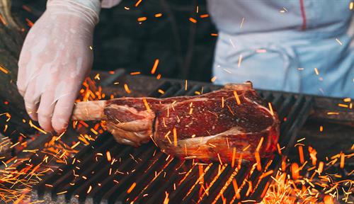 A cook grills a large, thick-cut beef steak on a charcoal fire.