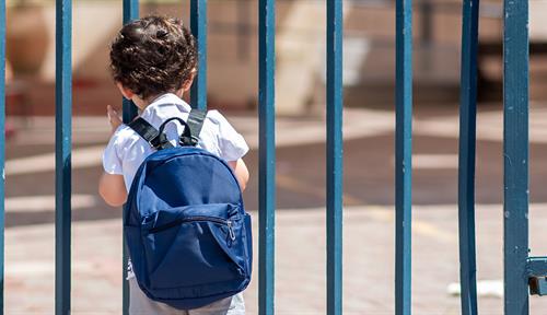 Photograph of a small child with a backpack staring through some railings at an empty schoolyard.