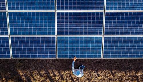  Photograph of a man seen from above. He is standing on grass and has his arm stretched out toward a wall of solar panels.